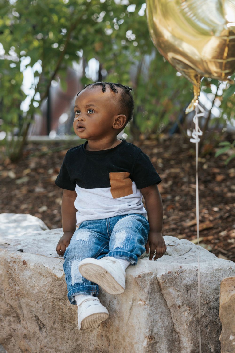 A child in casual wear sits on a stone wall with a gold balloon visible in the background.