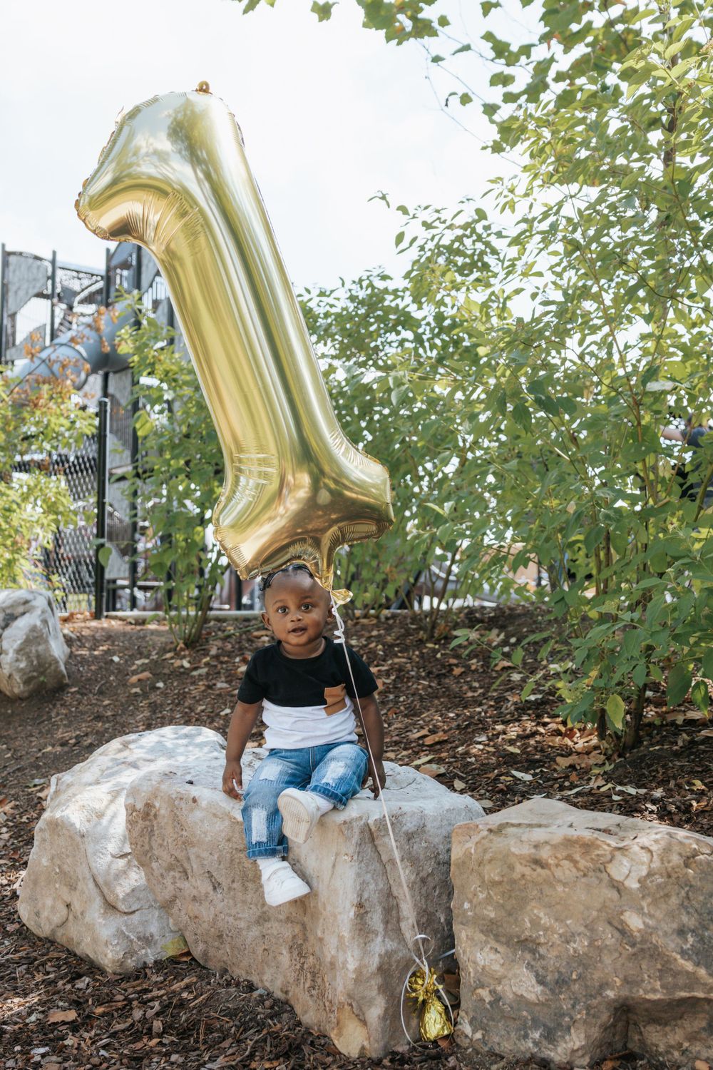 A child sits on a rock with a large gold number 1 balloon floating above in an outdoor garden setting.