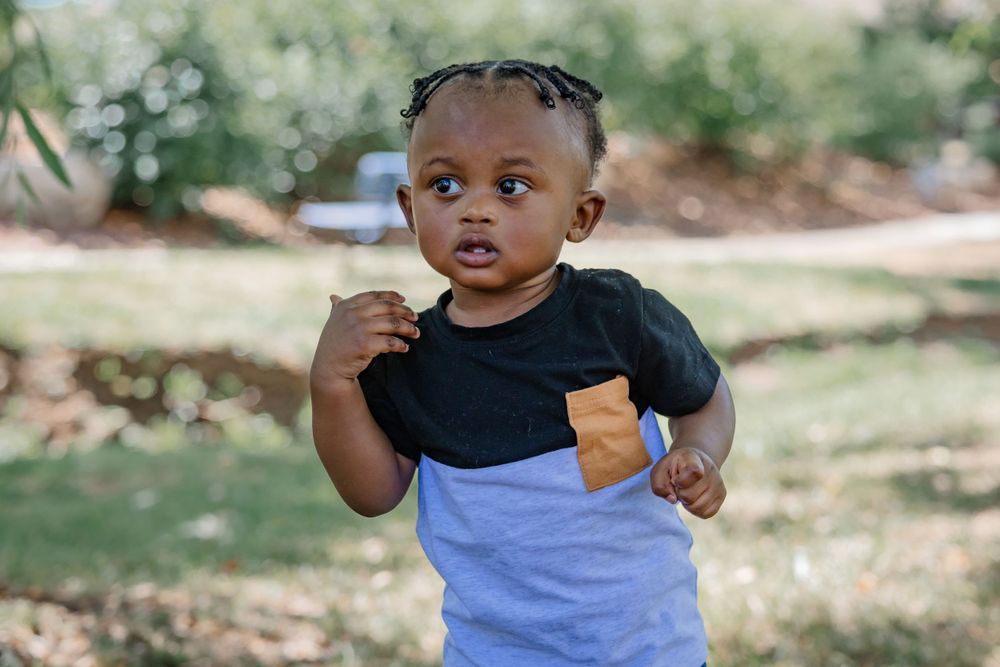 A toddler in a black and white shirt with brown pocket detail stands in an outdoor setting.