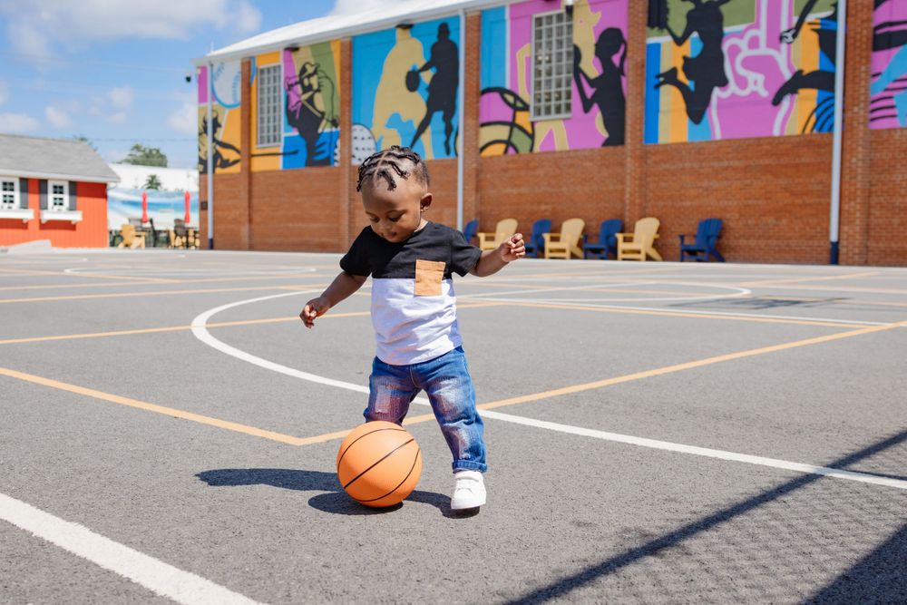 A young child dribbles a basketball on an outdoor court with colorful mural art in the background.