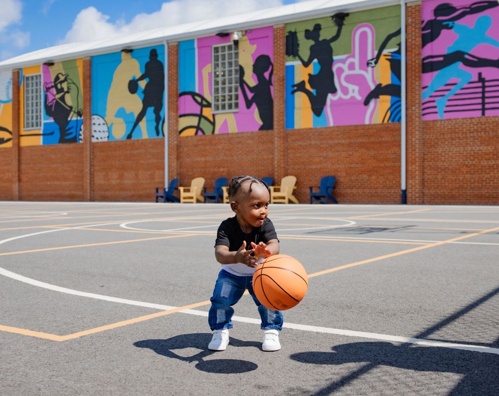 A child plays with a basketball on a court featuring vibrant wall murals and geometric line markings.