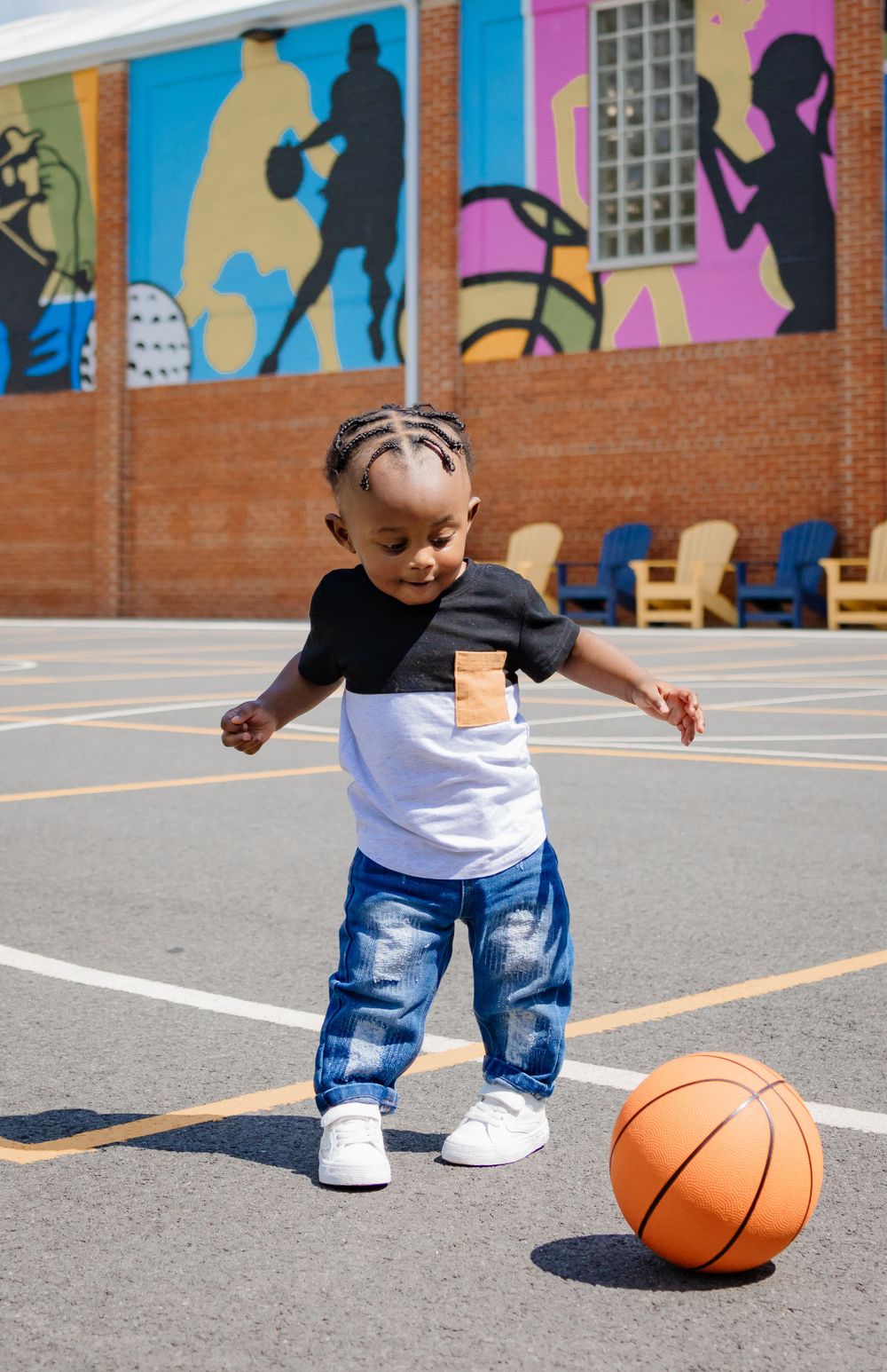 Young toddler playing with an orange basketball on a court with colorful sports murals in the background.