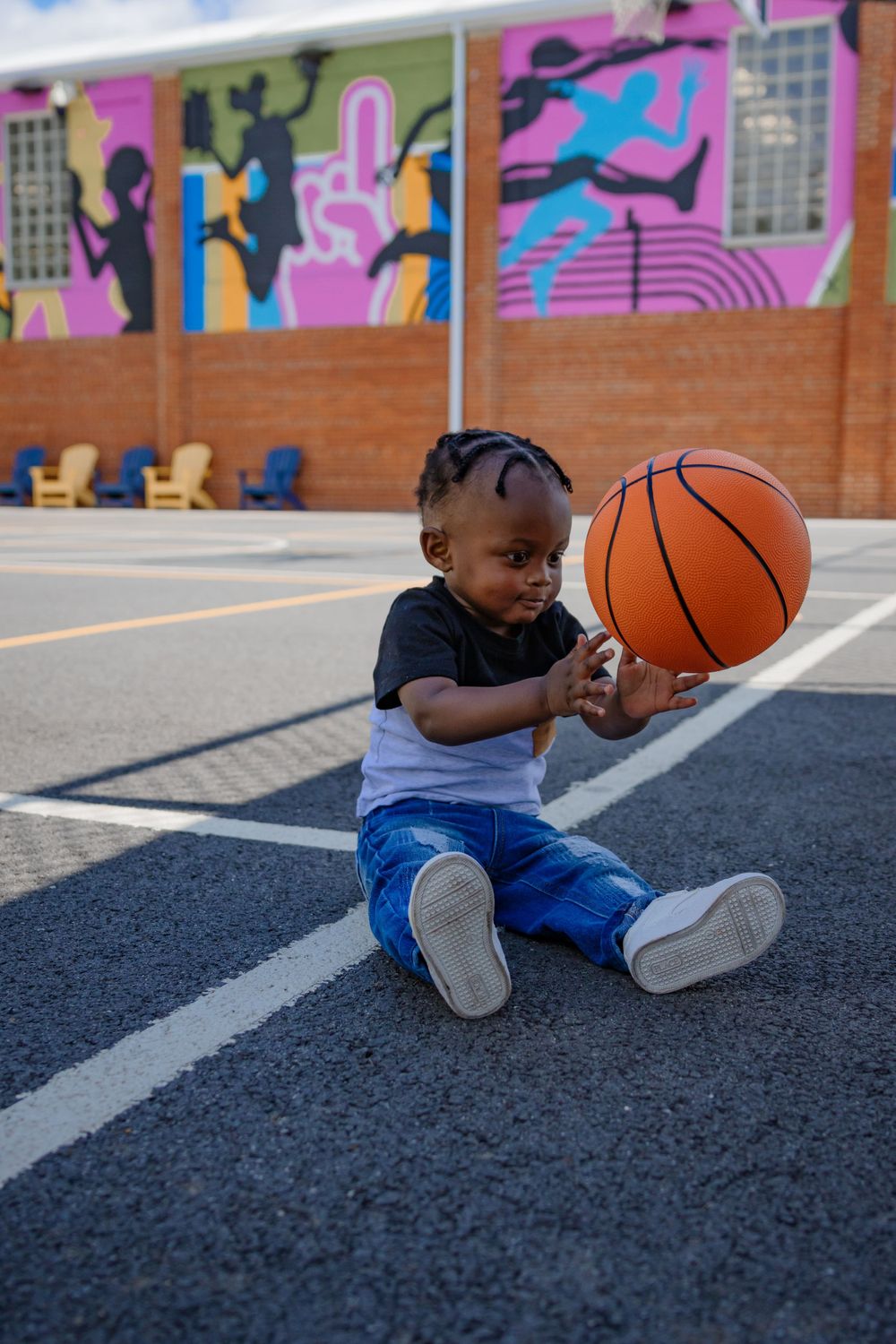 A small child plays with a basketball on a court with colorful murals in the background.