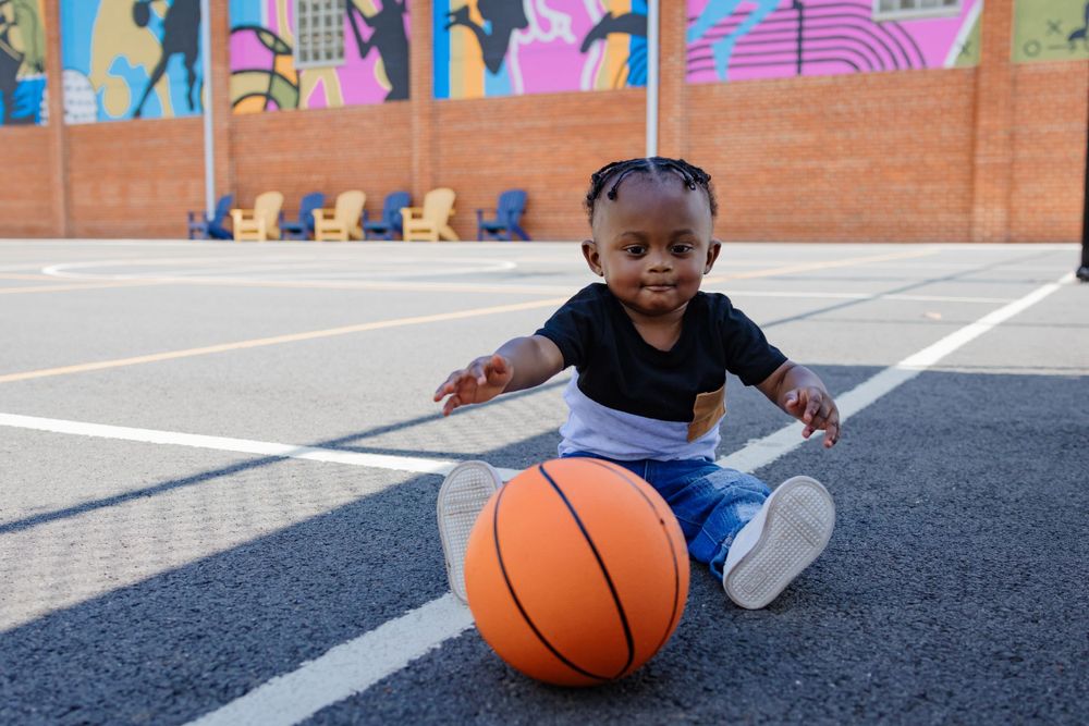 A toddler in casual wear sits on a basketball court with a ball and colorful wall art behind them.