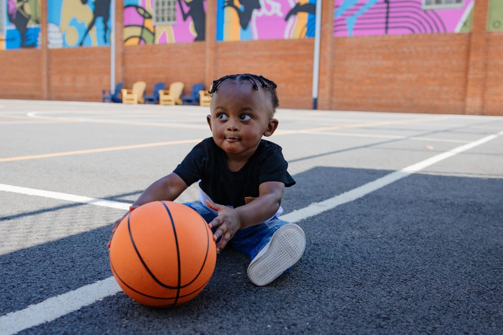 Small child sitting on basketball court with orange ball and colorful mural wall.