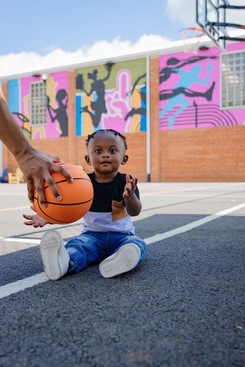 Toddler reaching for basketball while sitting on outdoor court with decorated wall.