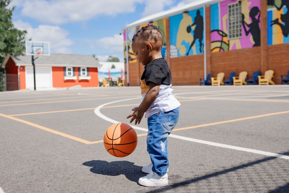 Child dribbling basketball on outdoor court with colorful sports-themed wall art.