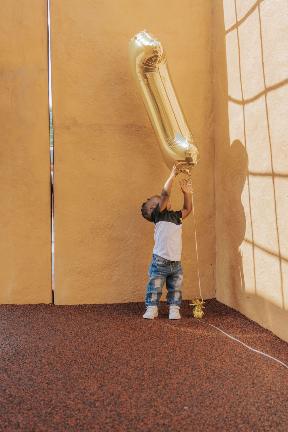 Small child reaching for gold number one balloon against orange wall.