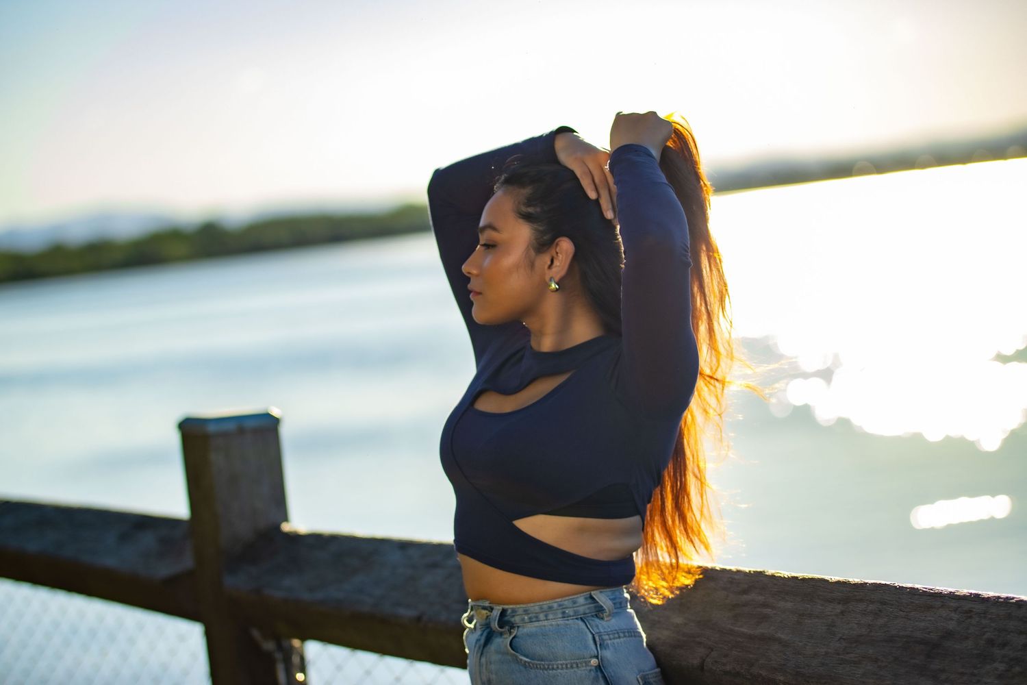 Silhouette at sunset by water, wearing crop top and jeans with long flowing hair.