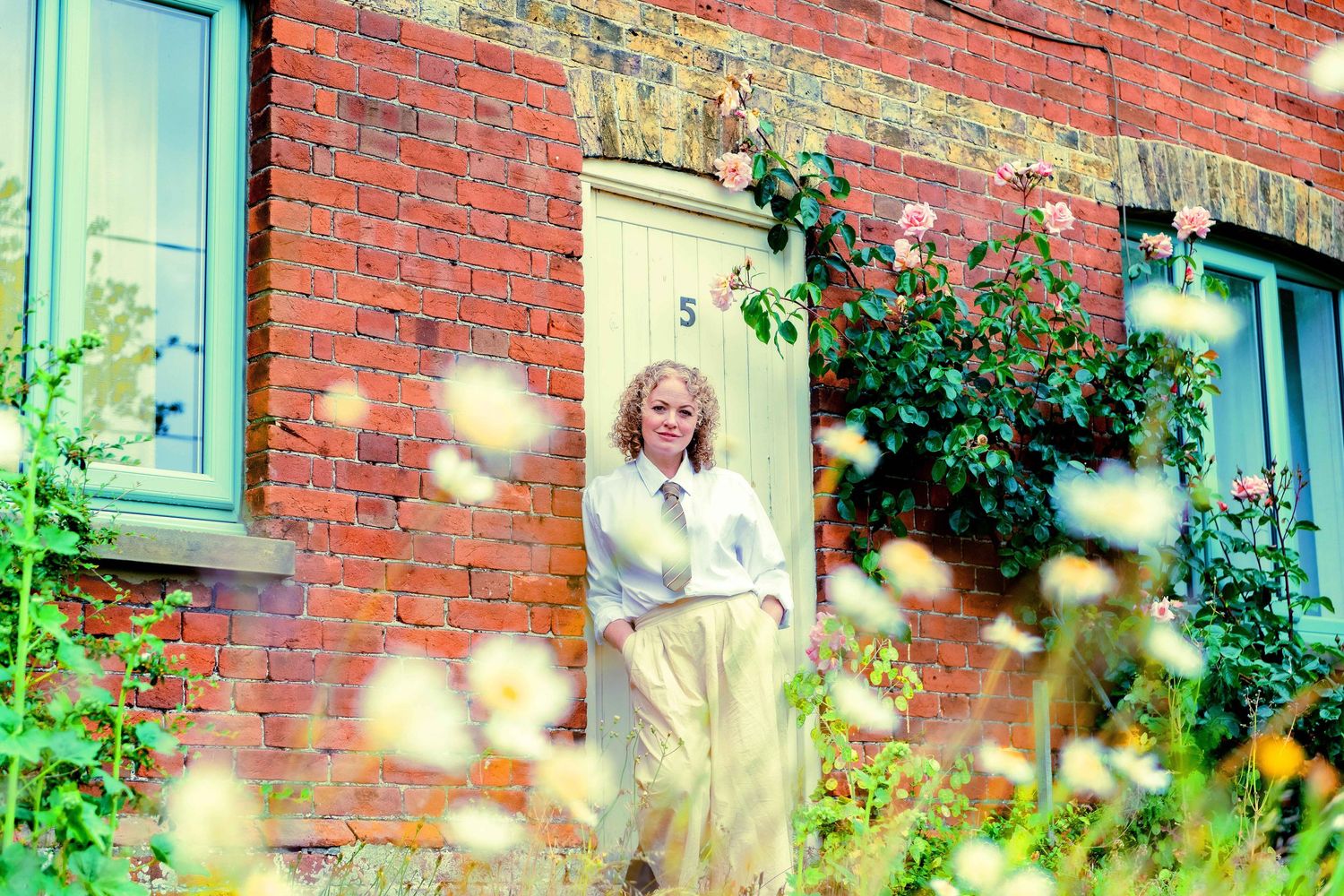A person in white stands by a yellow door of a red brick house surrounded by blooming flowers.