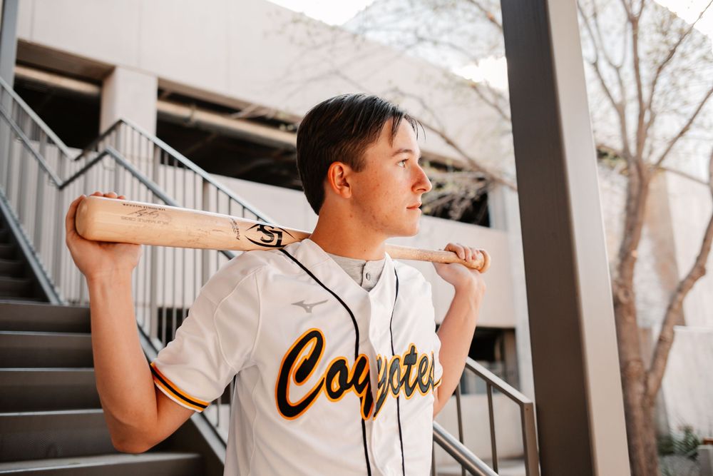 Baseball player in Coyotes uniform poses with bat resting on shoulders outdoors.