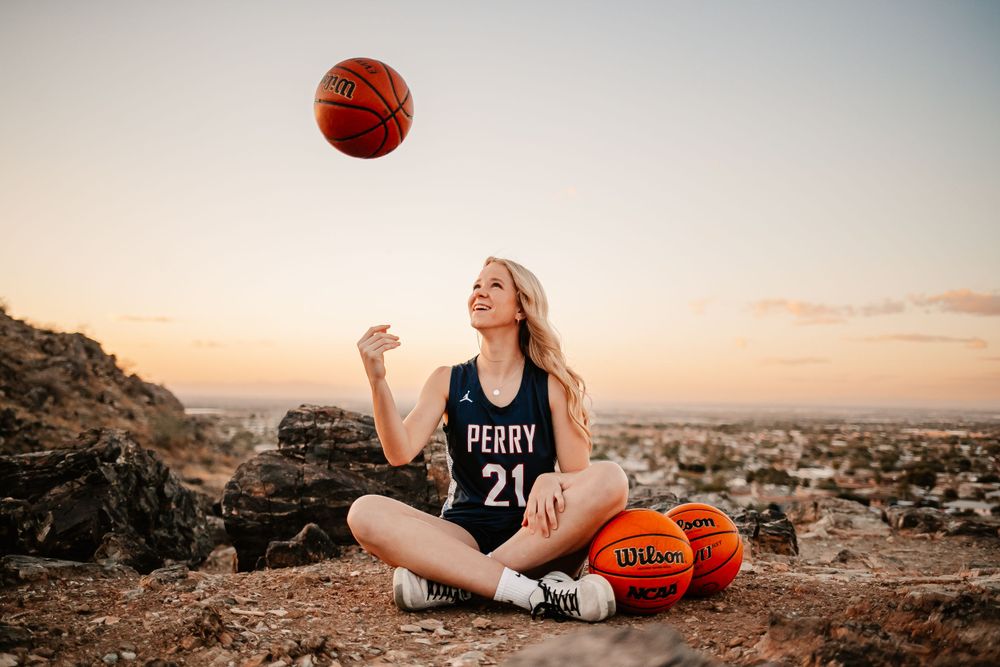 Blonde female basketball player in Perry number 21 jersey tossing ball at scenic sunset overlook.