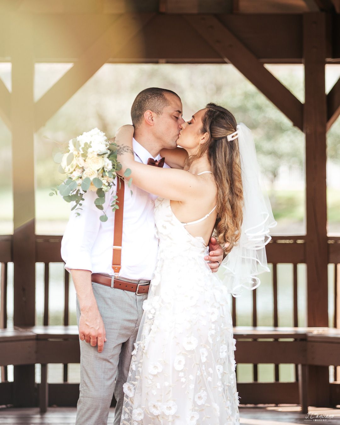 A romantic wedding couple shares a kiss under a wooden gazebo with the bride holding a white floral bouquet.