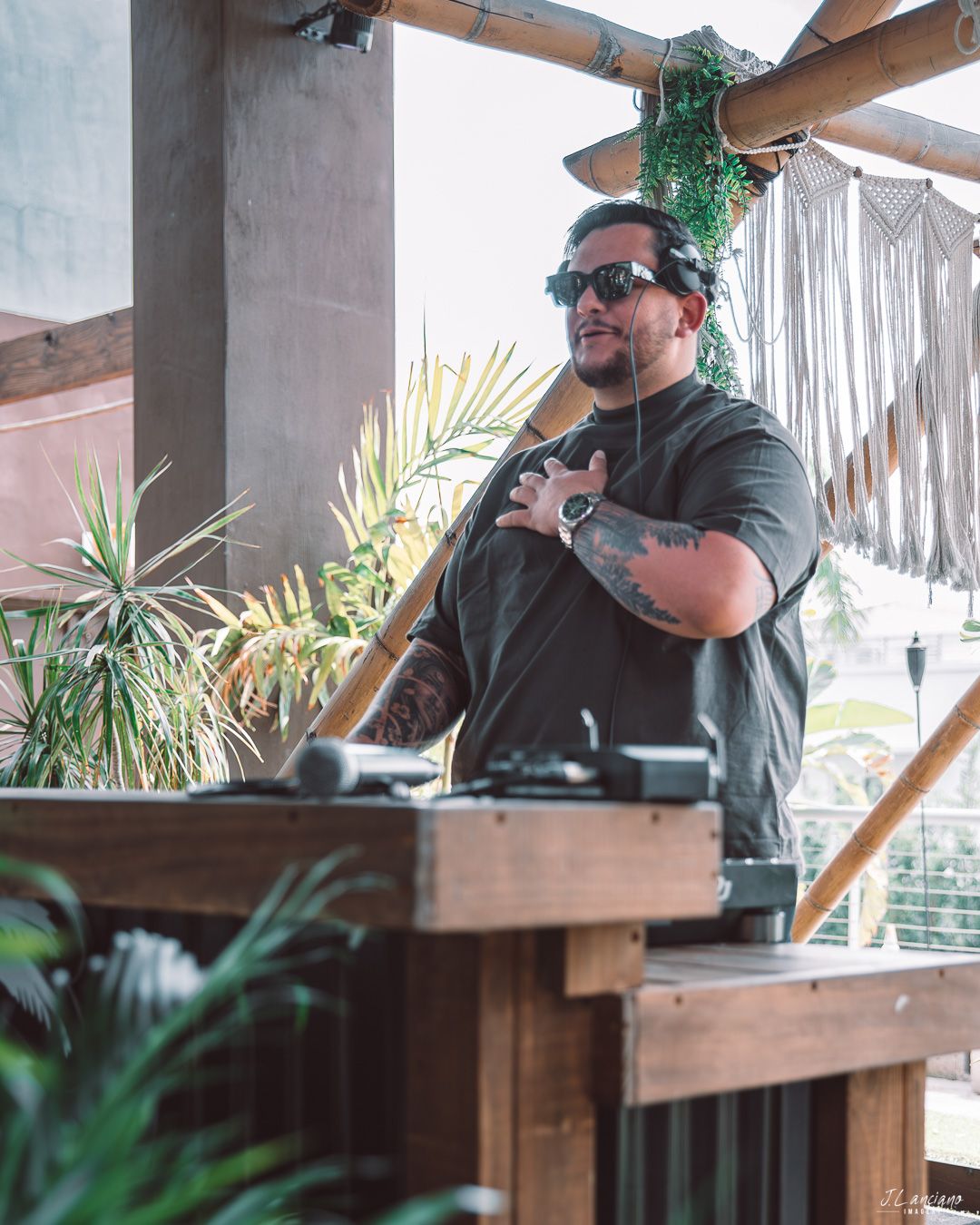 DJ performing outdoors at tropical venue with palm plants and wooden deck setup.