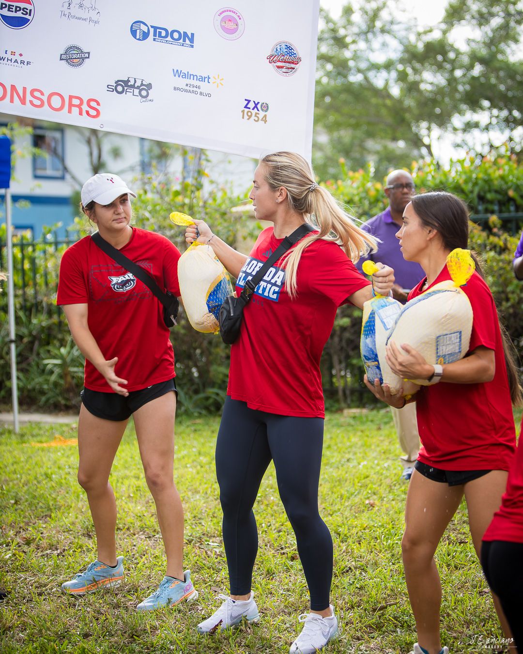 Three people in red shirts holding packages during an outdoor charitable event under a sponsor banner.
