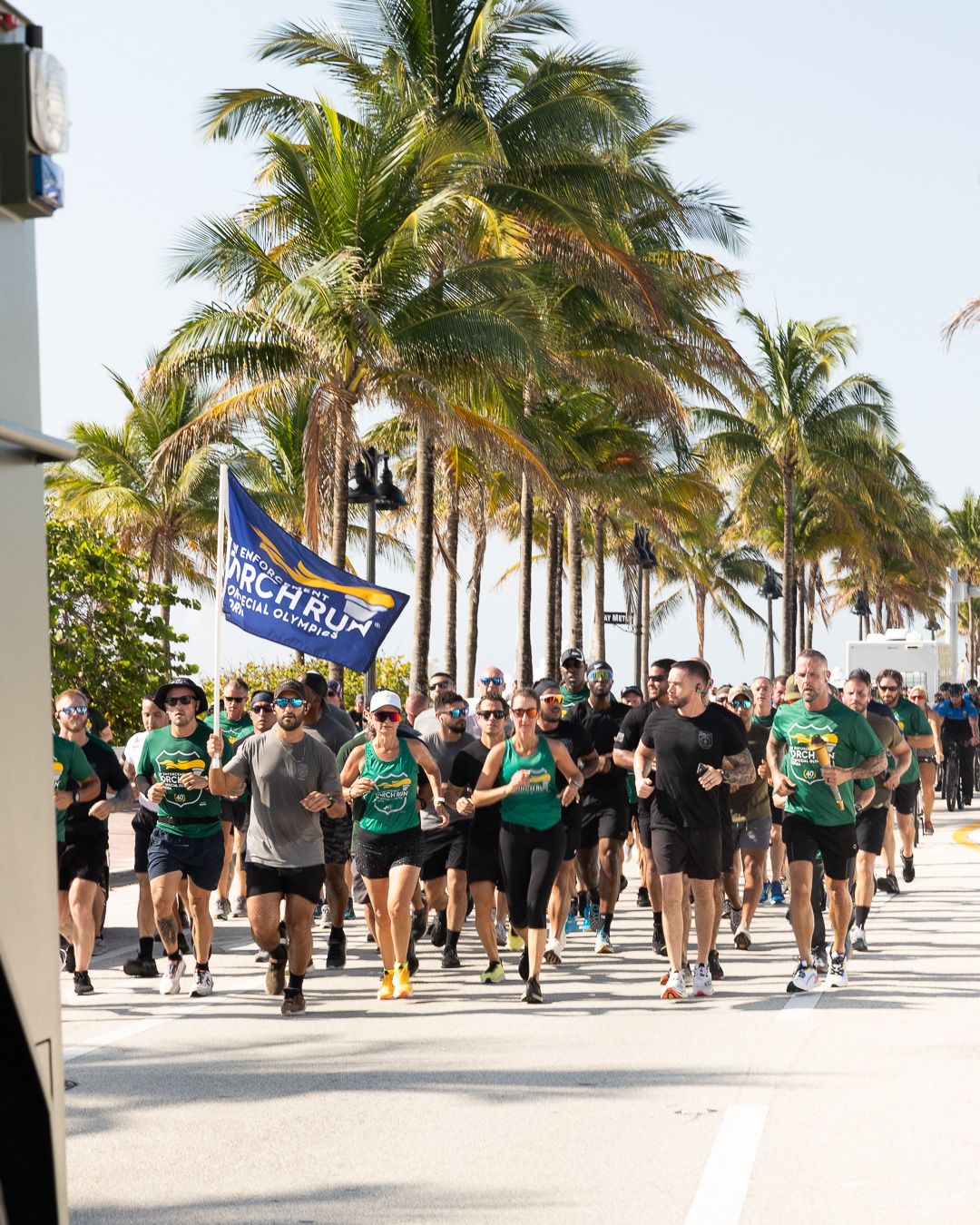 Large group of runners in green shirts jogging along a palm tree-lined beachfront path with a flag.