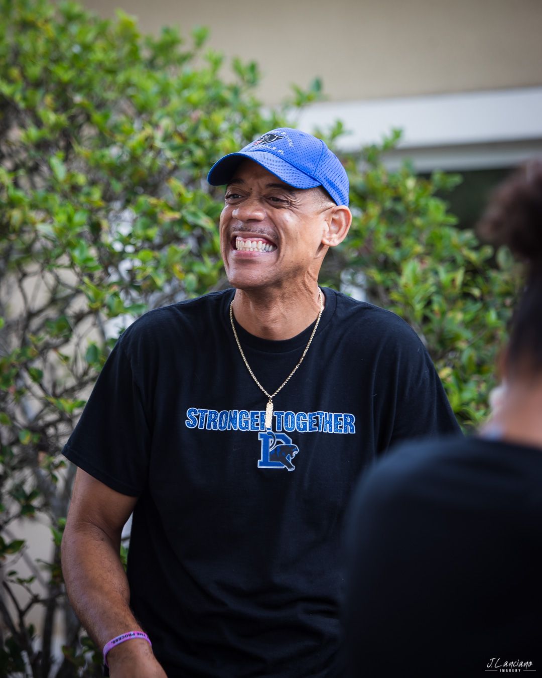 Person wearing a black t-shirt and blue baseball cap smiling outdoors with greenery in background.