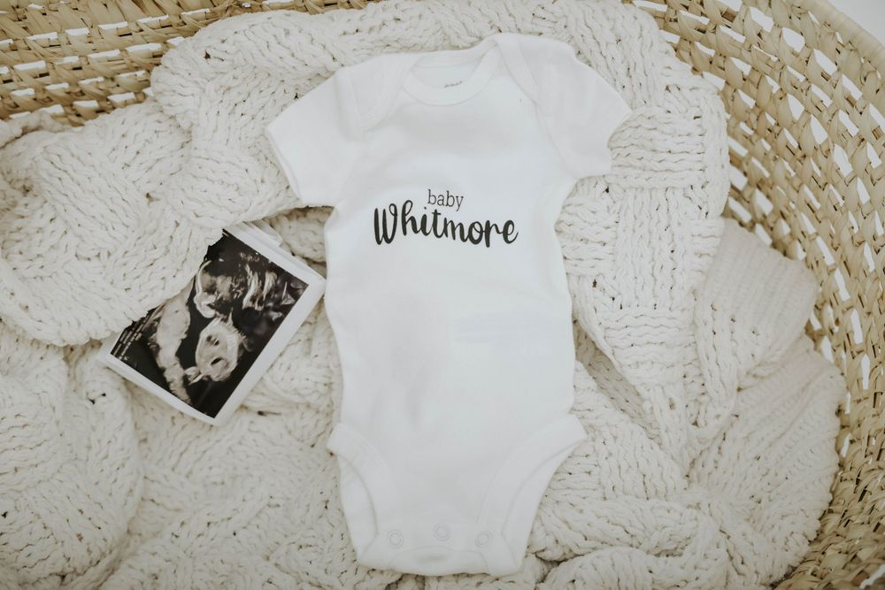 White baby onesie with 'Whitmore' text and ultrasound photo on knit blanket.