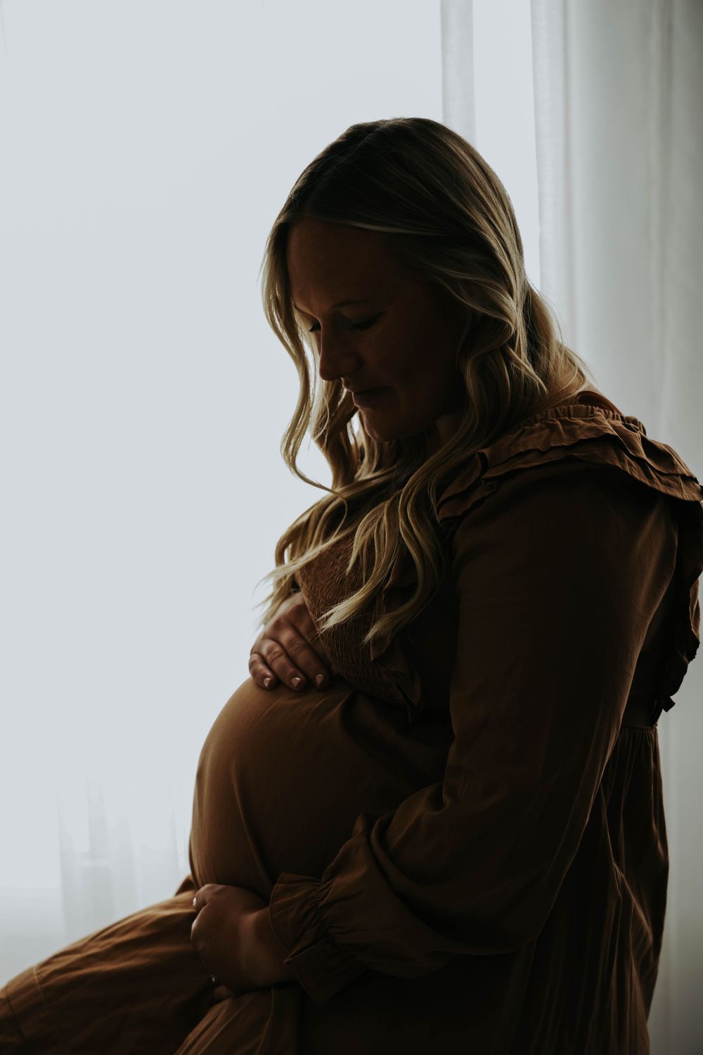 Silhouette of pregnant figure in brown dress by window.