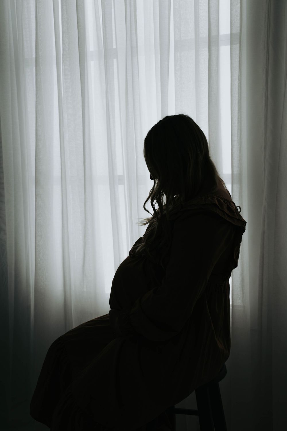 Dramatic silhouette of pregnant person sitting by window with sheer curtains.