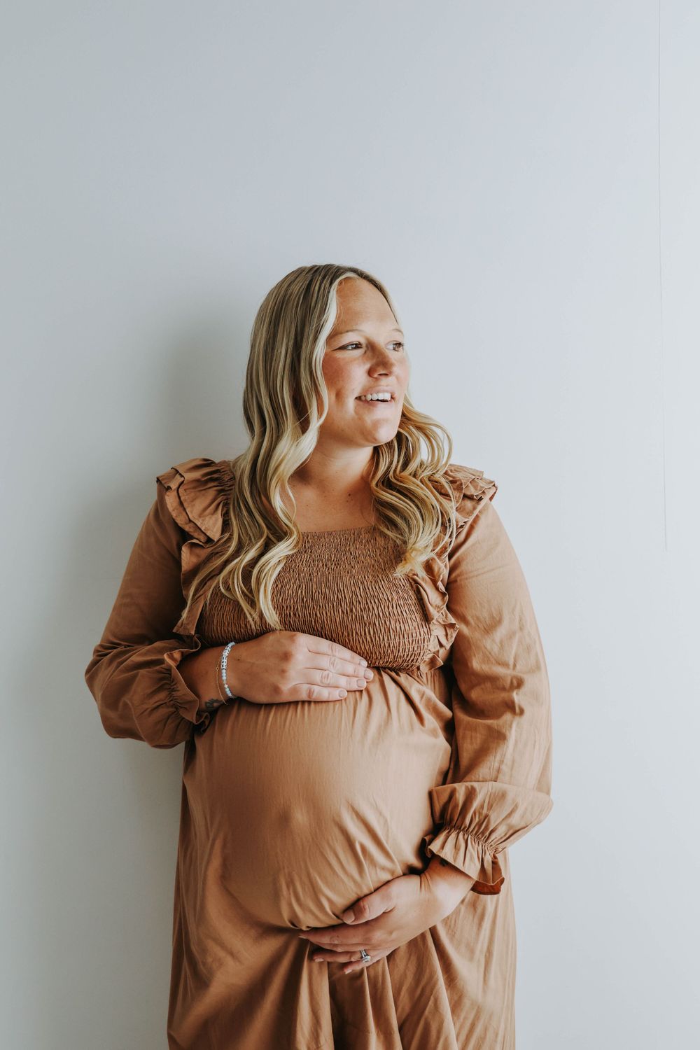 Expectant mother wearing brown long-sleeved dress stands against white wall with gentle smile.