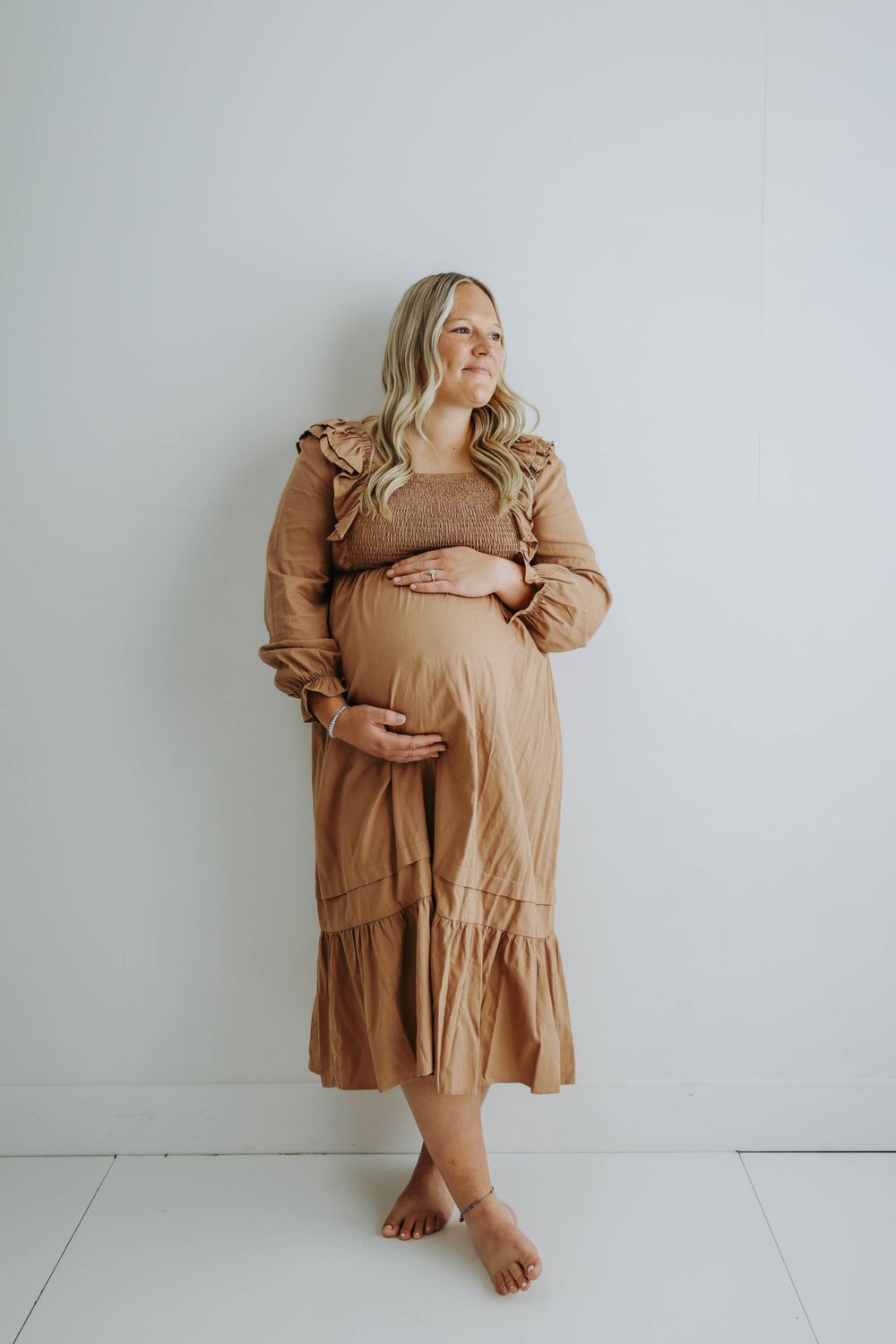 Pregnant woman in flowing caramel dress poses barefoot against minimalist white background.