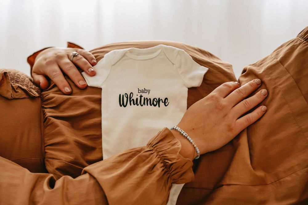 Close-up of hands holding white baby onesie with Whitmore text against brown fabric.