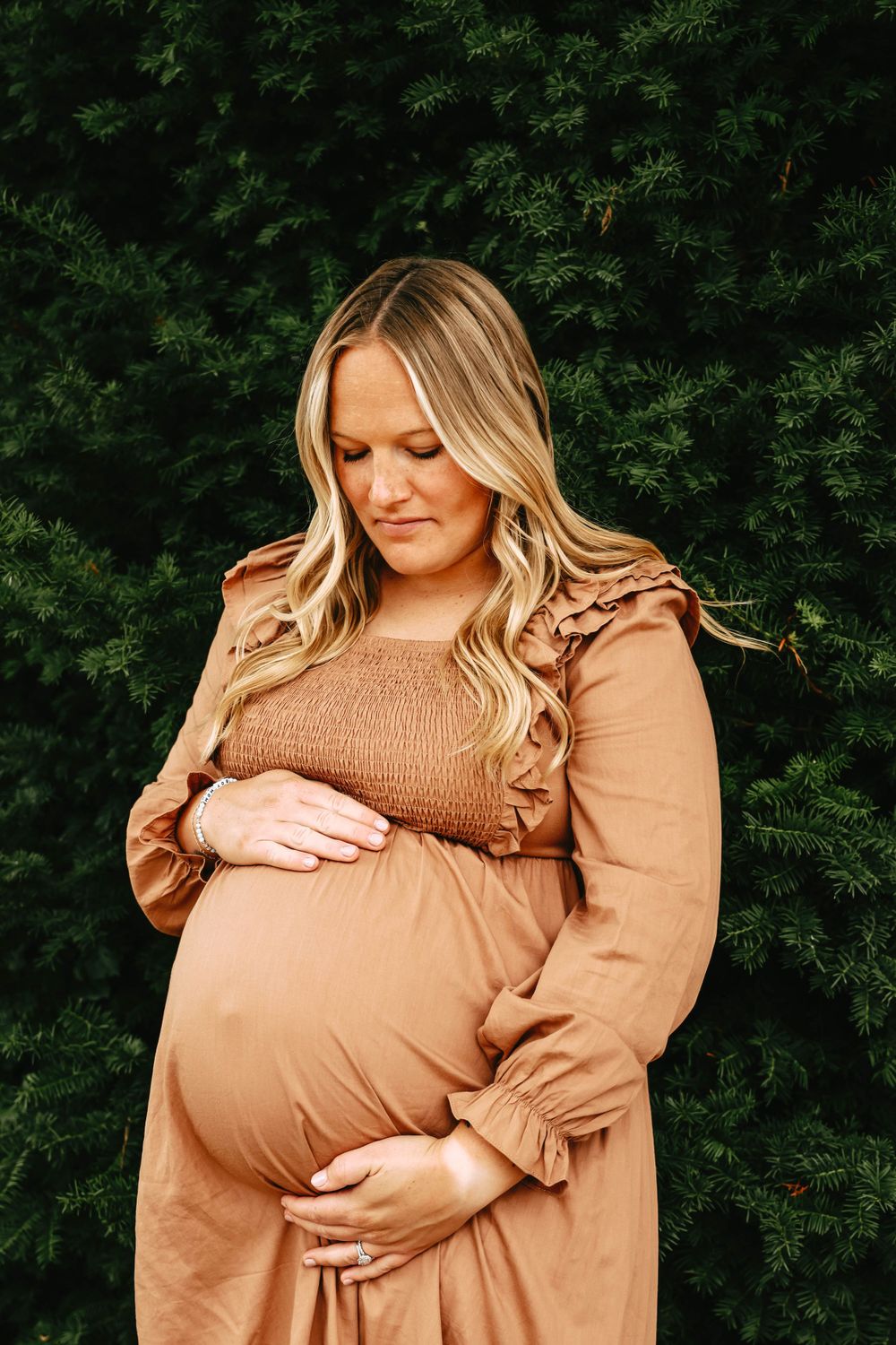Maternity portrait against lush green pine trees wearing brown ruffle dress.