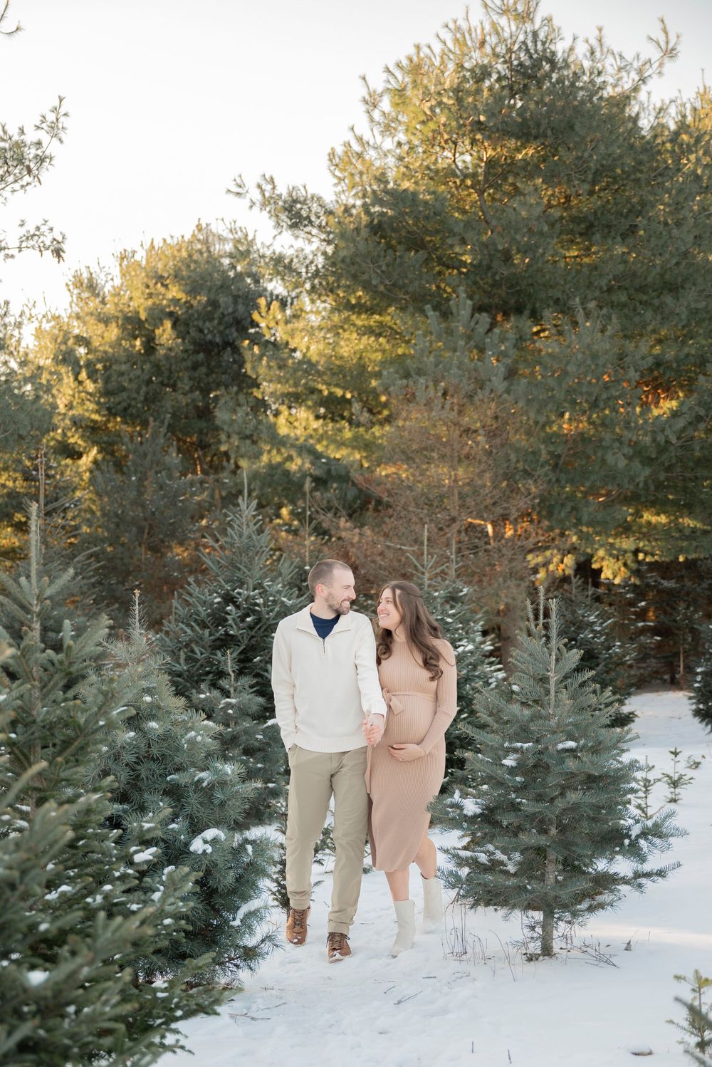 A pregnant couple walks among snow-covered evergreen trees in a Christmas tree farm during golden hour in Lake Orion Michigan
