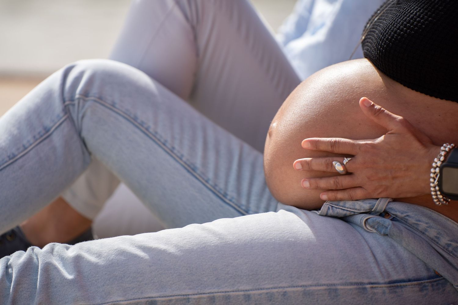 Close-up shot of pregnant belly and hands wearing jewelry against light denim background.