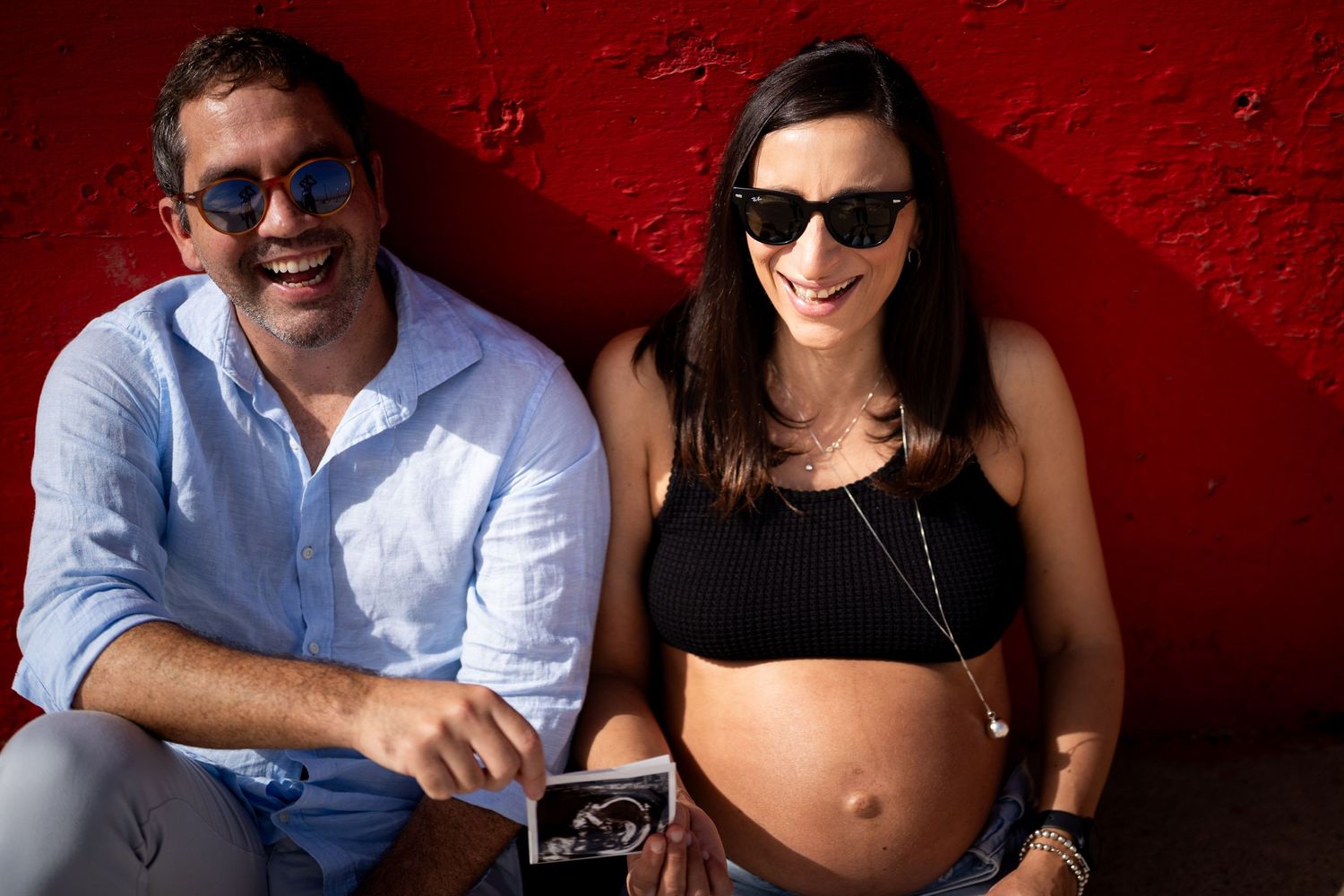 Happy couple sharing ultrasound photo during outdoor maternity session against red wall.