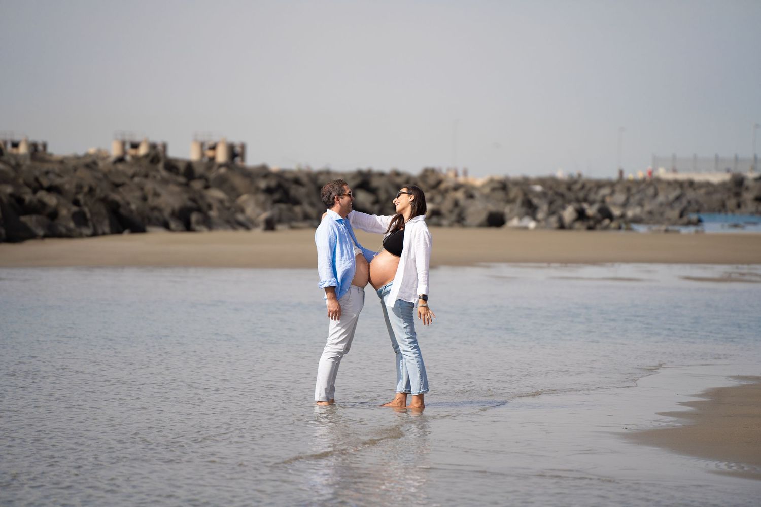 A couple stands together on a sandy beach with rocks and ocean in the background.