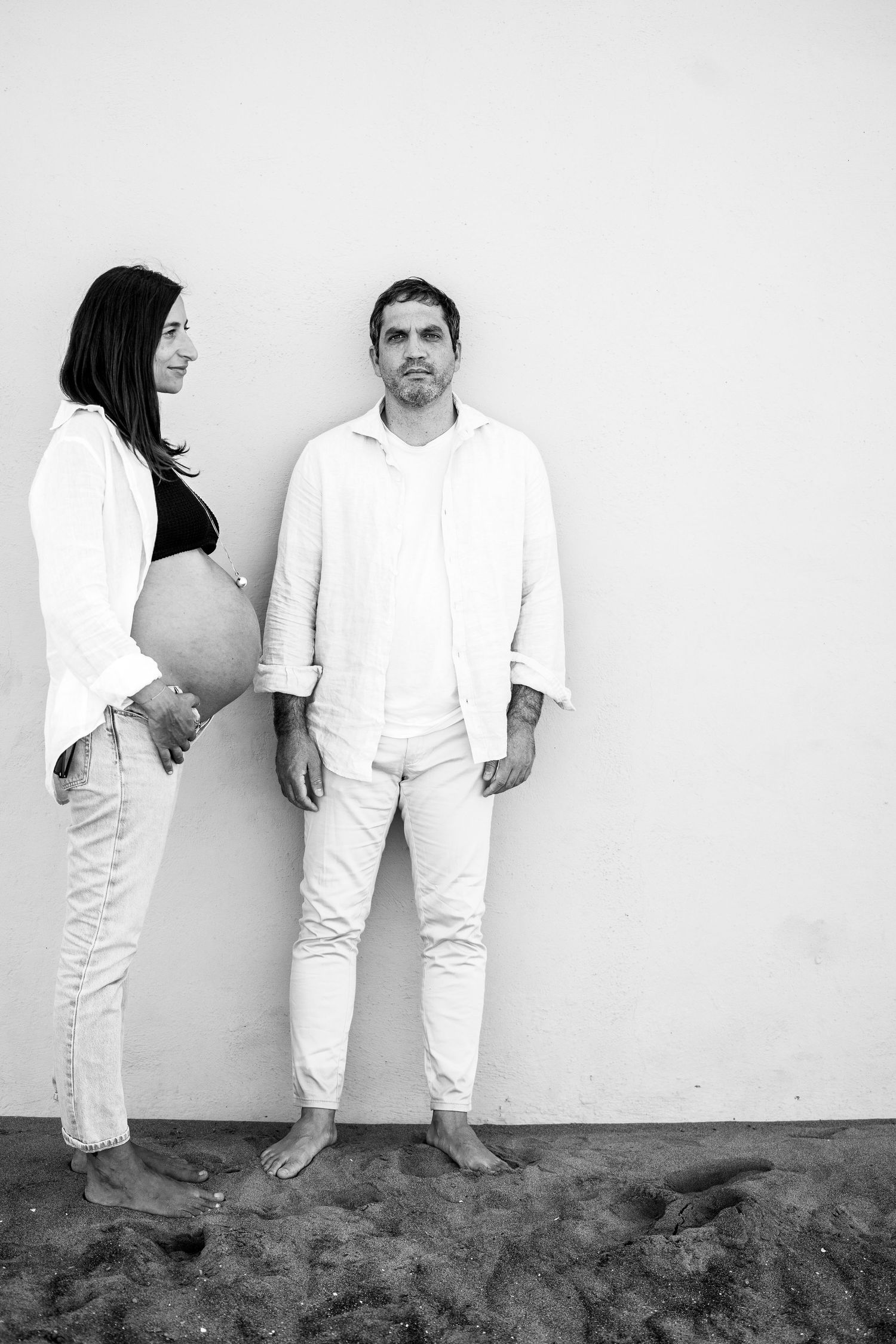 A couple in white clothing poses against a plain white wall.
