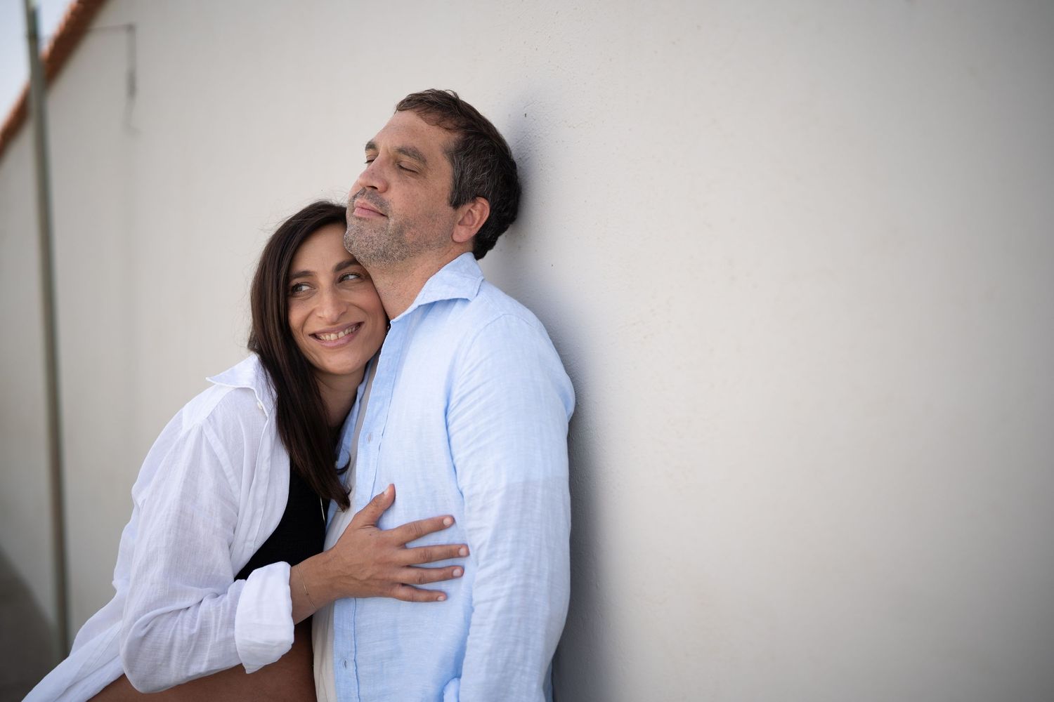 A couple embraces while leaning against a light colored wall.