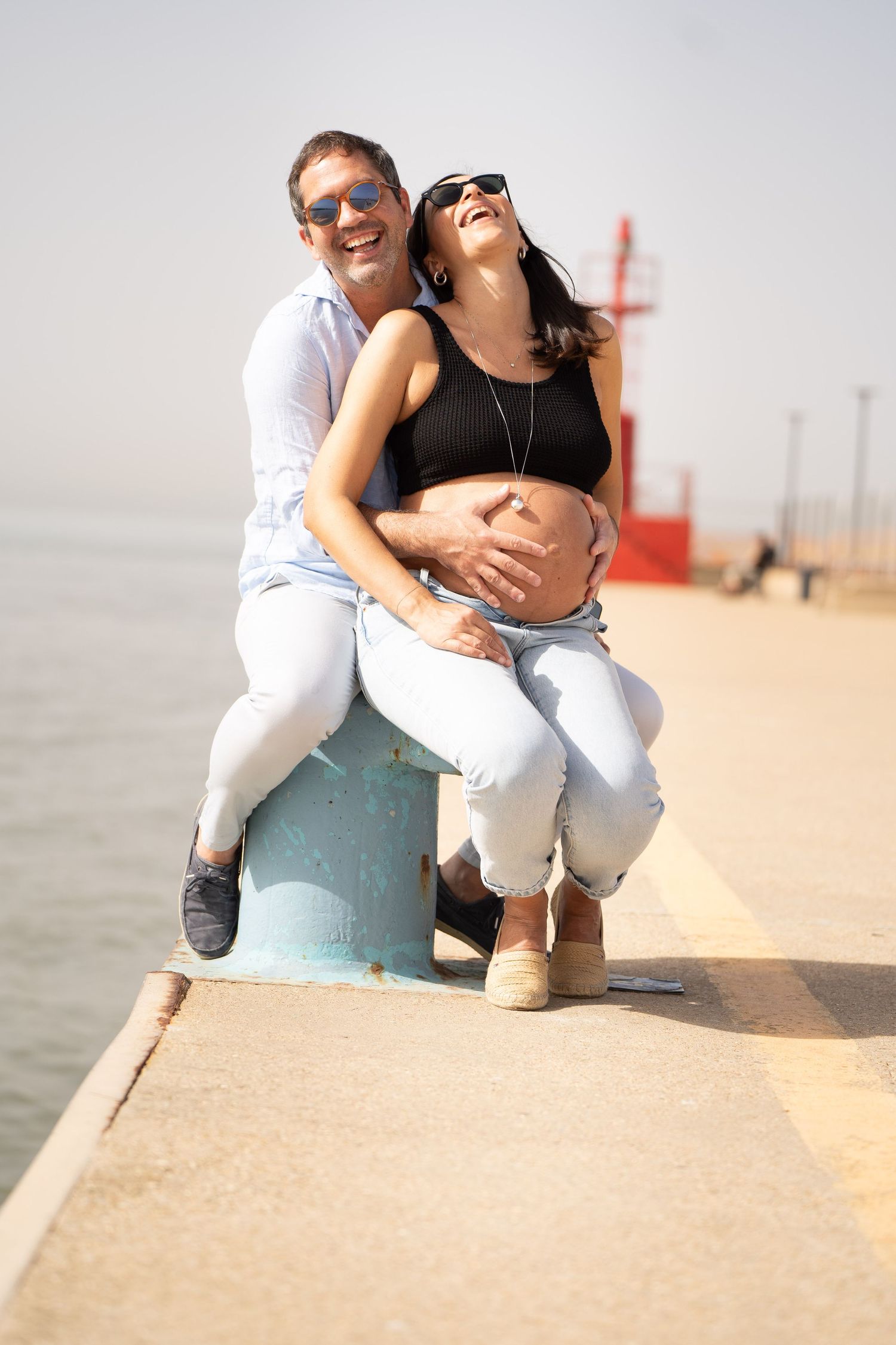 Expecting couple shares joyful moment on waterfront pier with red lighthouse in background.