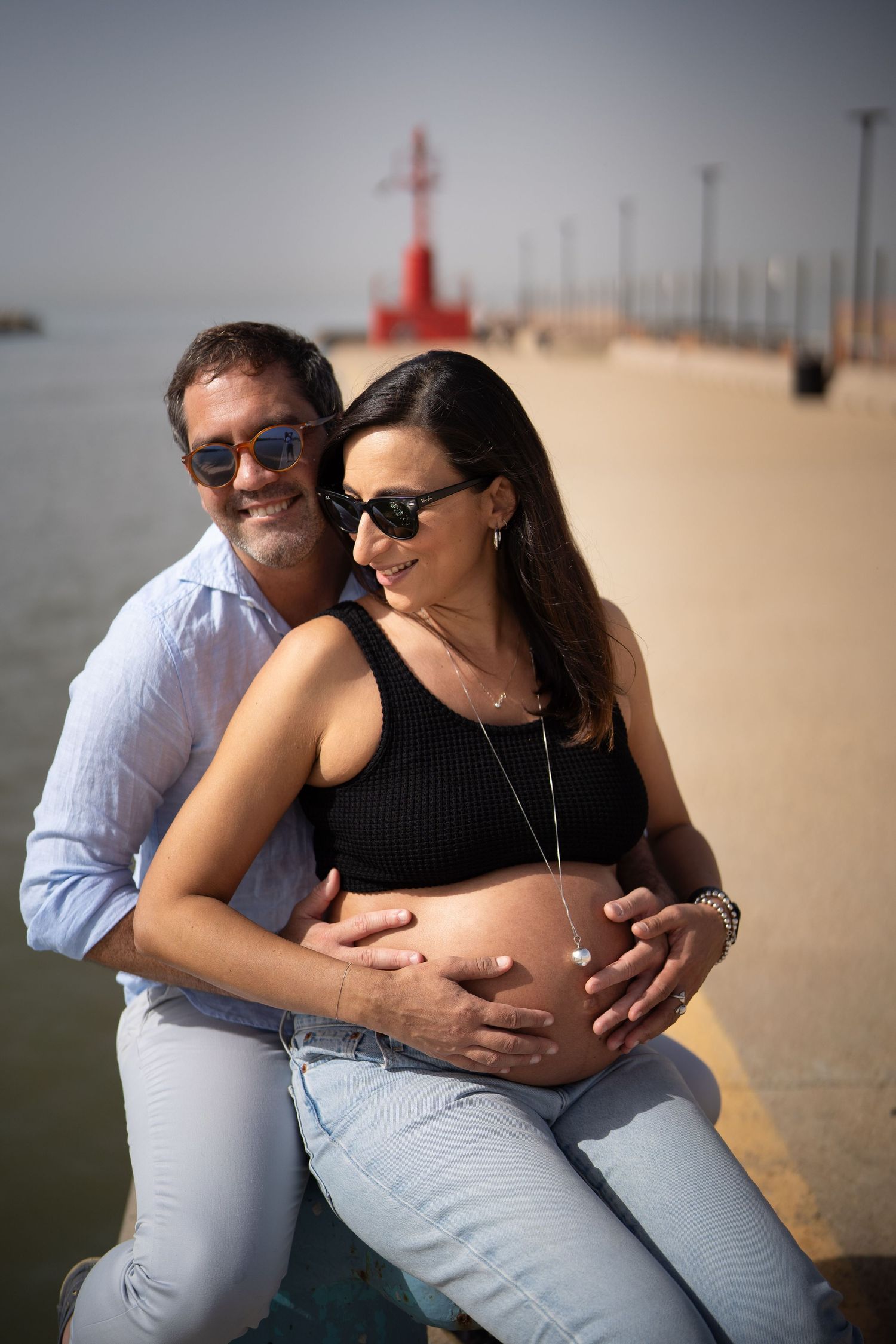Pregnancy photoshoot featuring couple sitting on pier with red lighthouse in background.