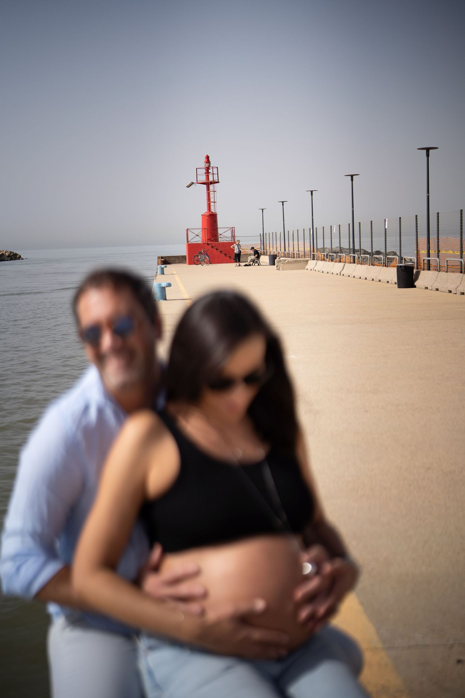 Artistic maternity photo with blurred lighthouse and pier creating dreamy atmosphere.