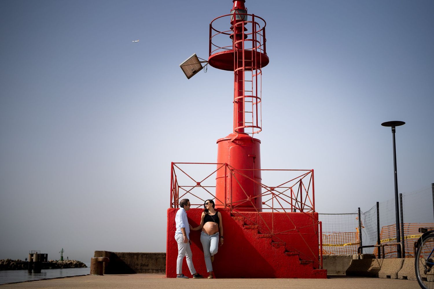 Couple poses against bright red lighthouse structure during maternity photoshoot.