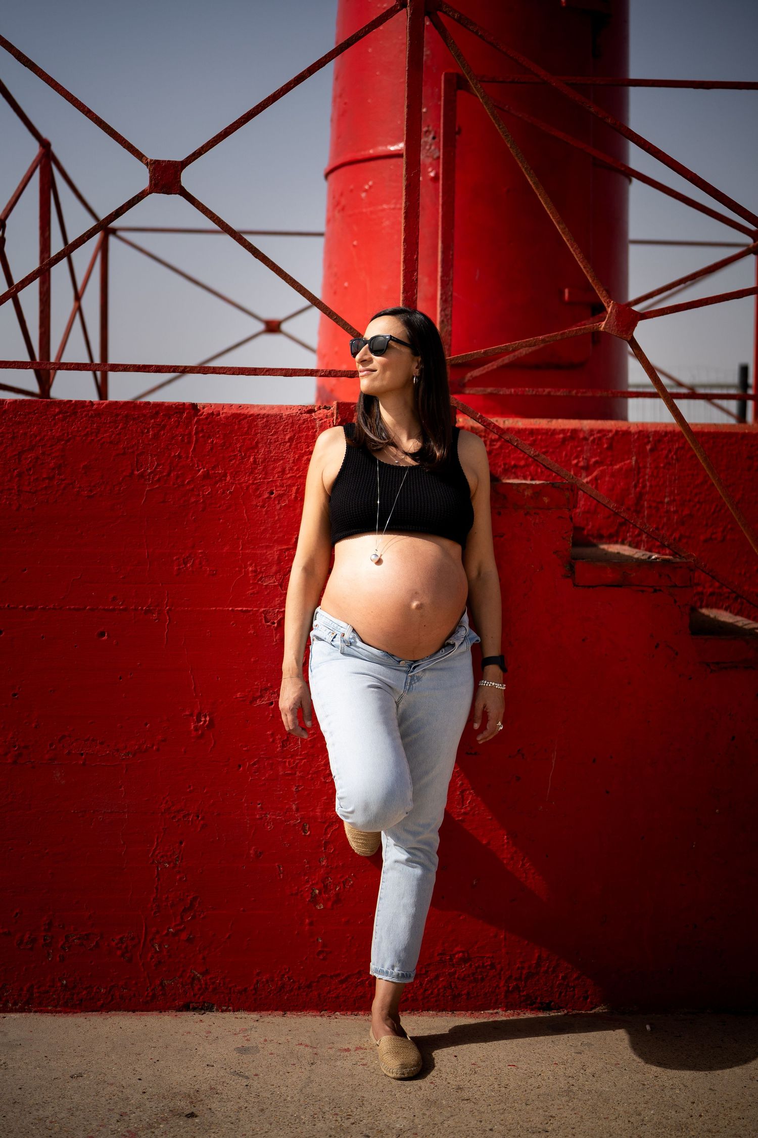 Maternity photo of expectant mother in black crop top and jeans posing against red lighthouse structure.