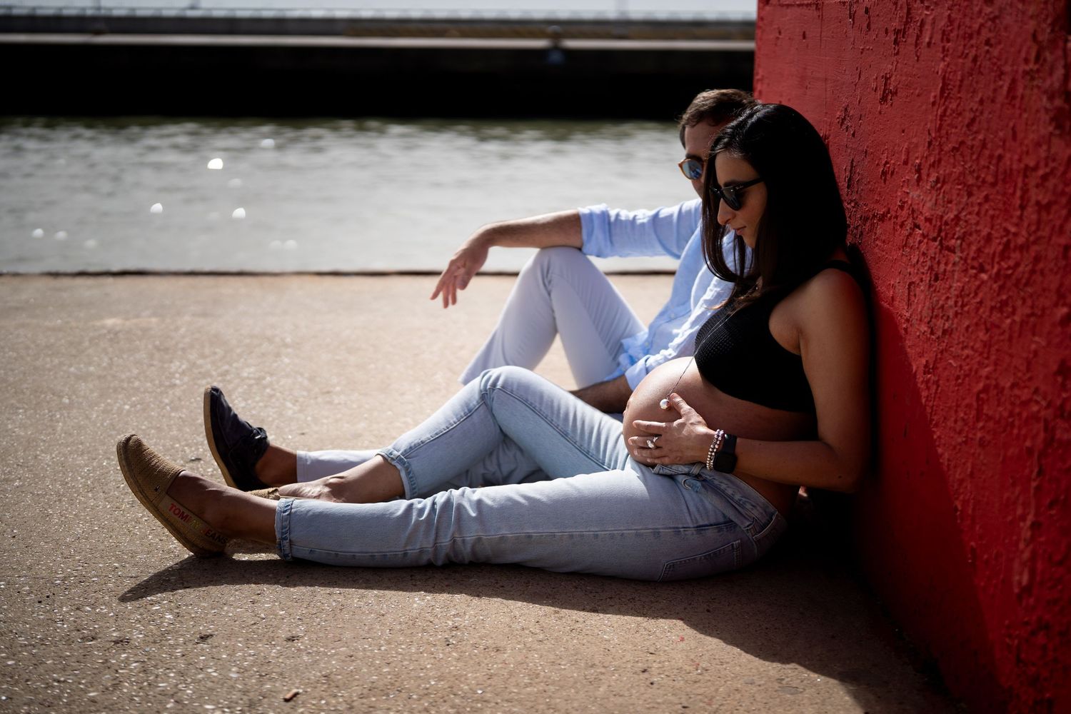 Maternity photo session with couple sitting by red wall near waterfront.