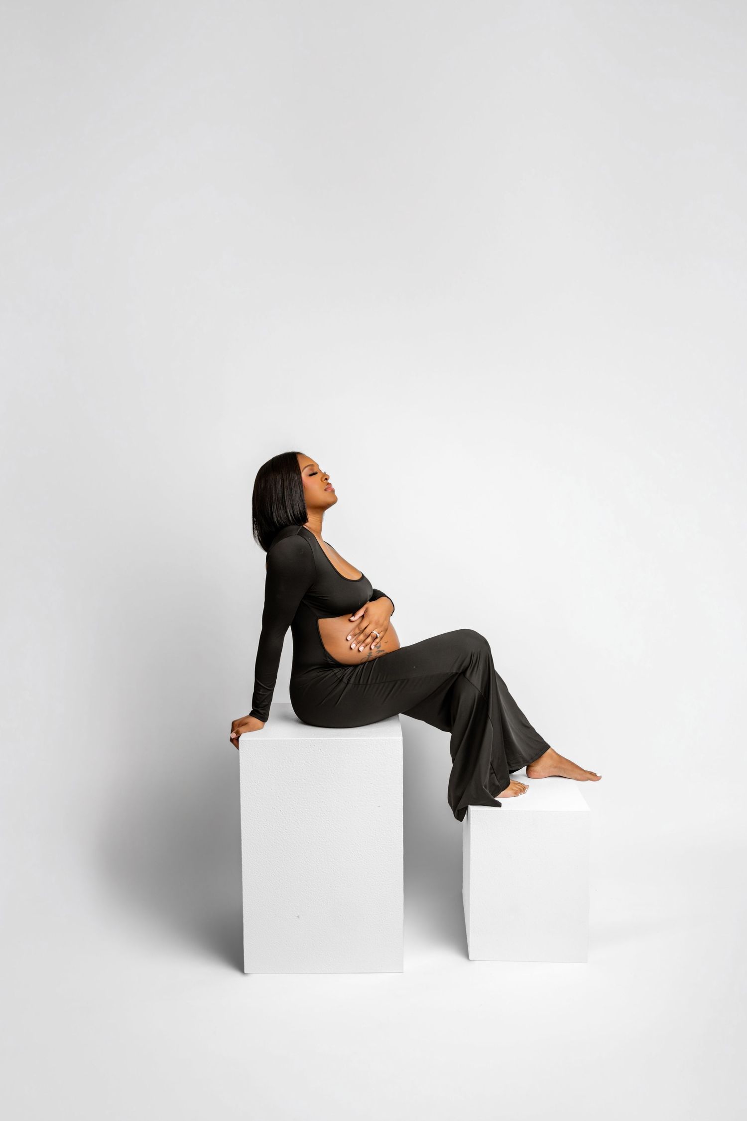 A woman in dark olive crop top and wide-leg pants sits gracefully on white cubes against a light background.