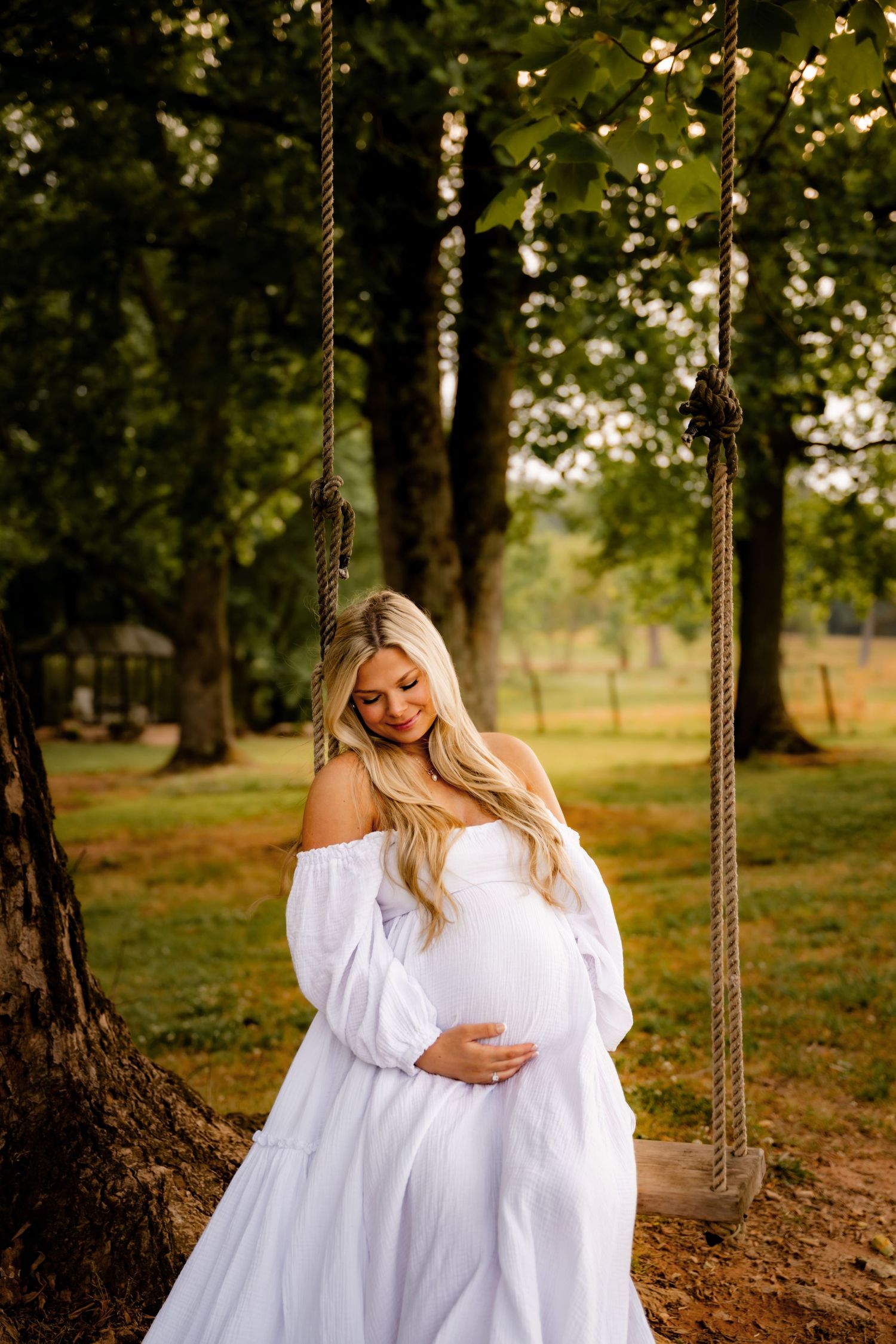 Pregnant woman in white off-shoulder dress cradling baby bump by rope swing in lush green park setting.