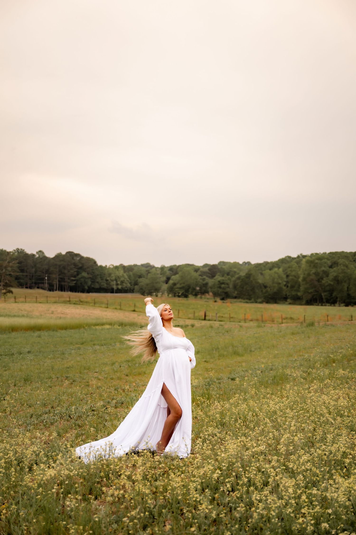 Pregnant woman in white flowing dress standing in a wildflower field, arms raised, hair blowing in the wind.