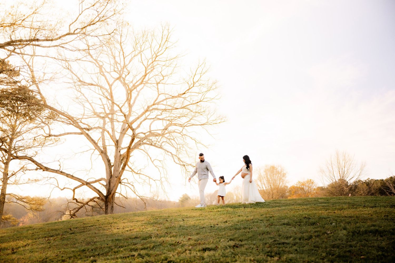 A couple in white outfits walks with their dog on a grassy hillside during golden hour beneath a bare tree.