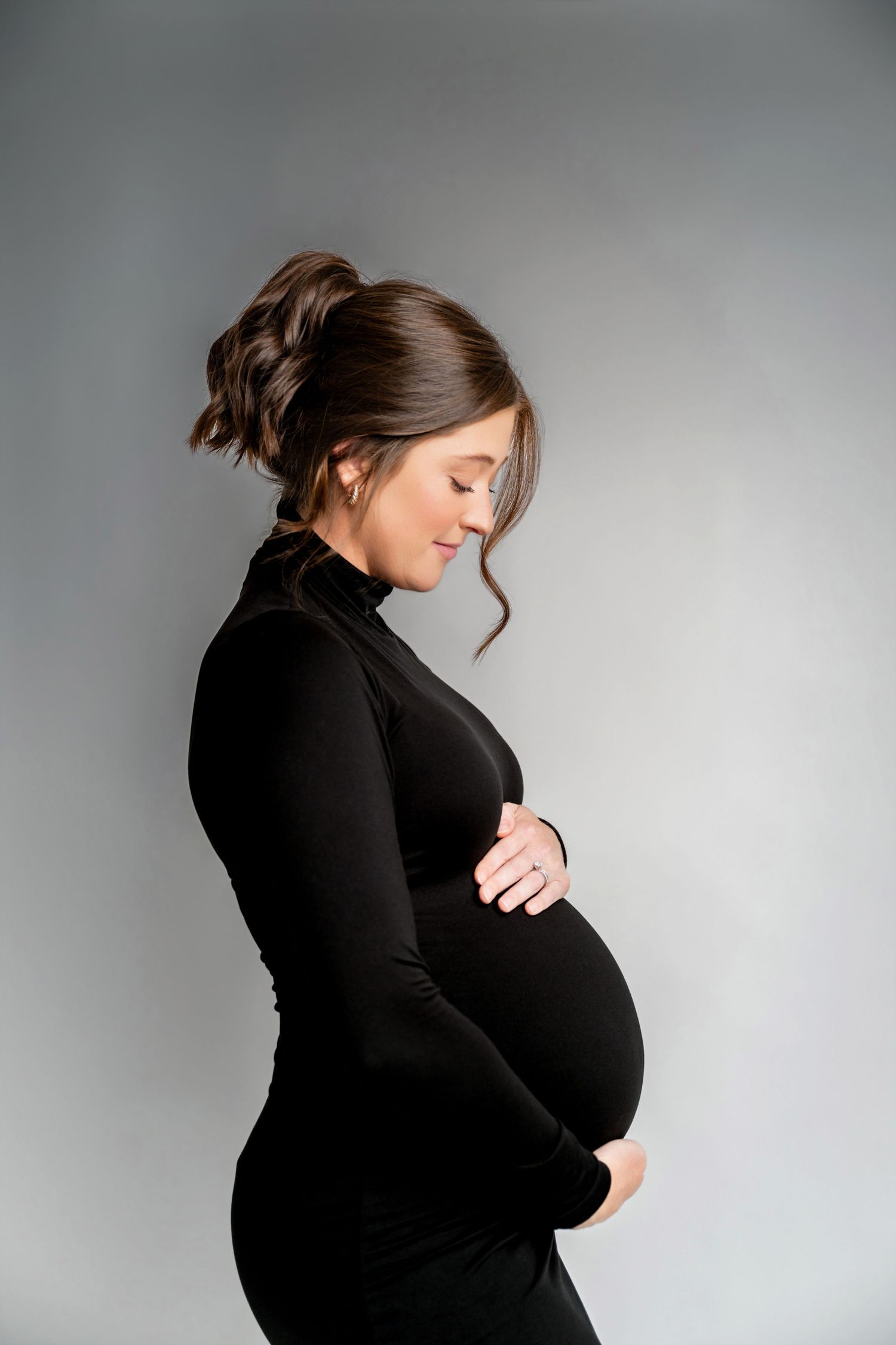 Pregnant woman in black dress cradling baby bump, studio maternity portrait on gray background.
