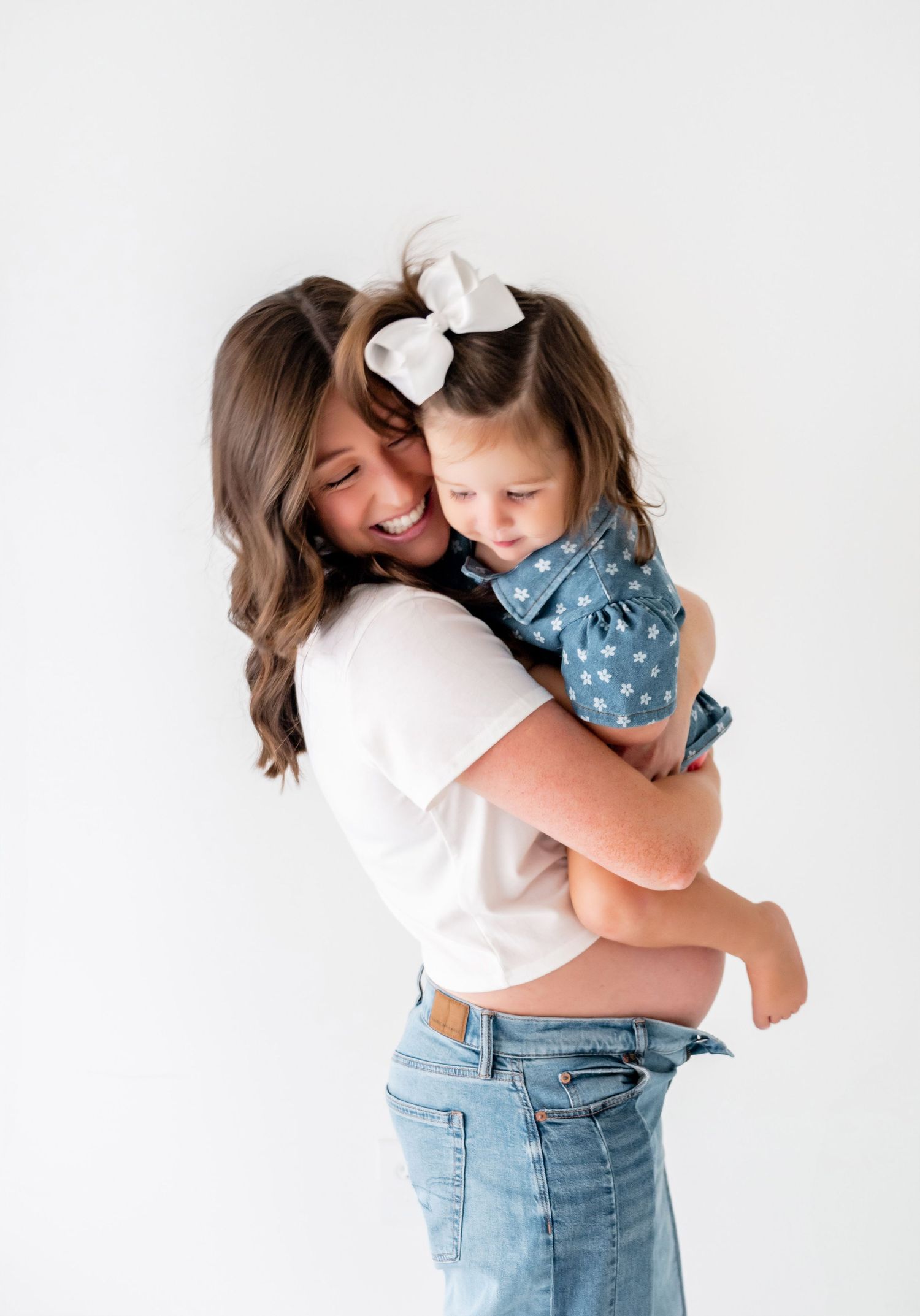 A smiling woman in a white tee and jeans hugs a toddler girl in a blue star dress with a white bow.