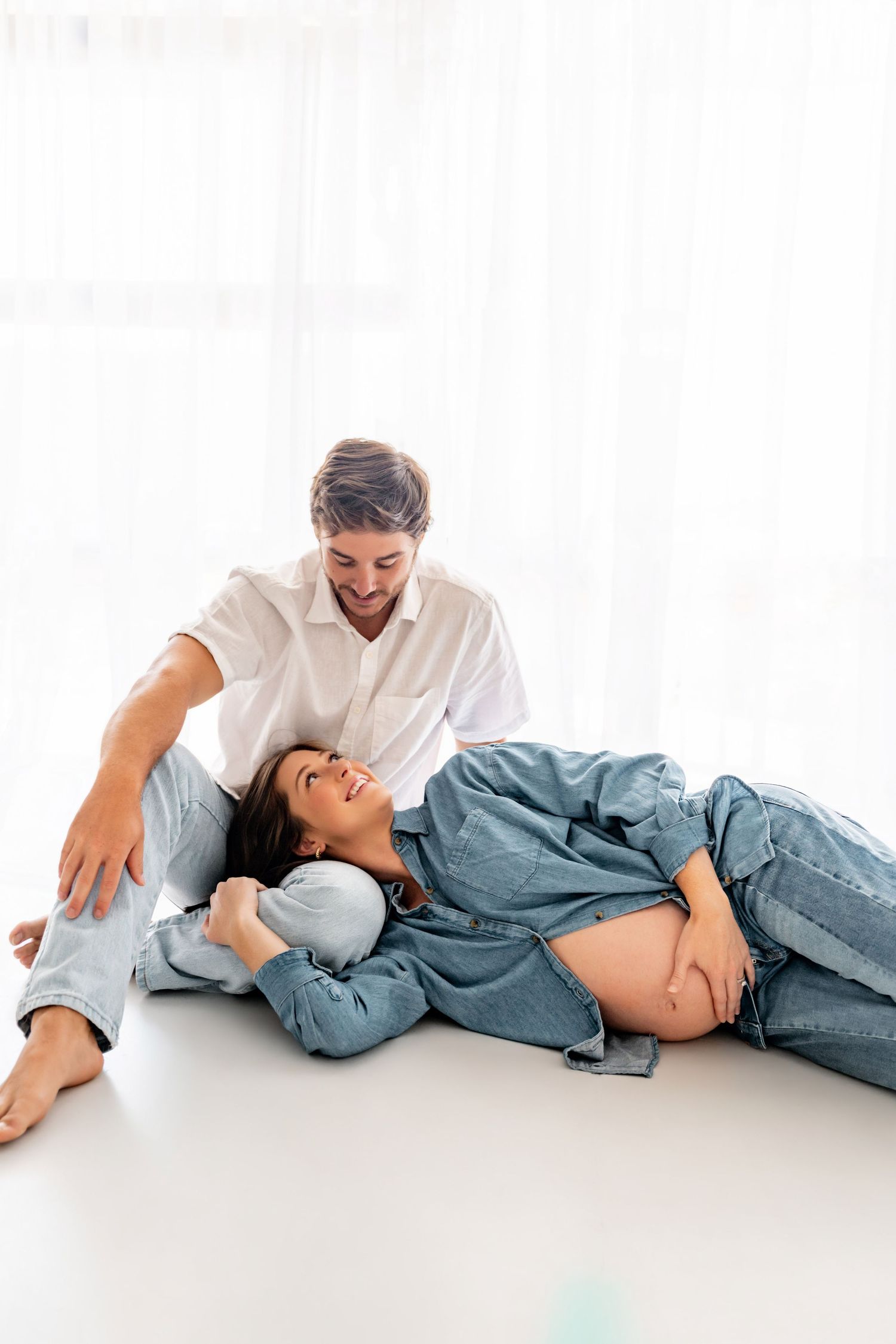 Happy couple expecting a baby, woman lying on man's lap touching her bare pregnant belly on white floor.