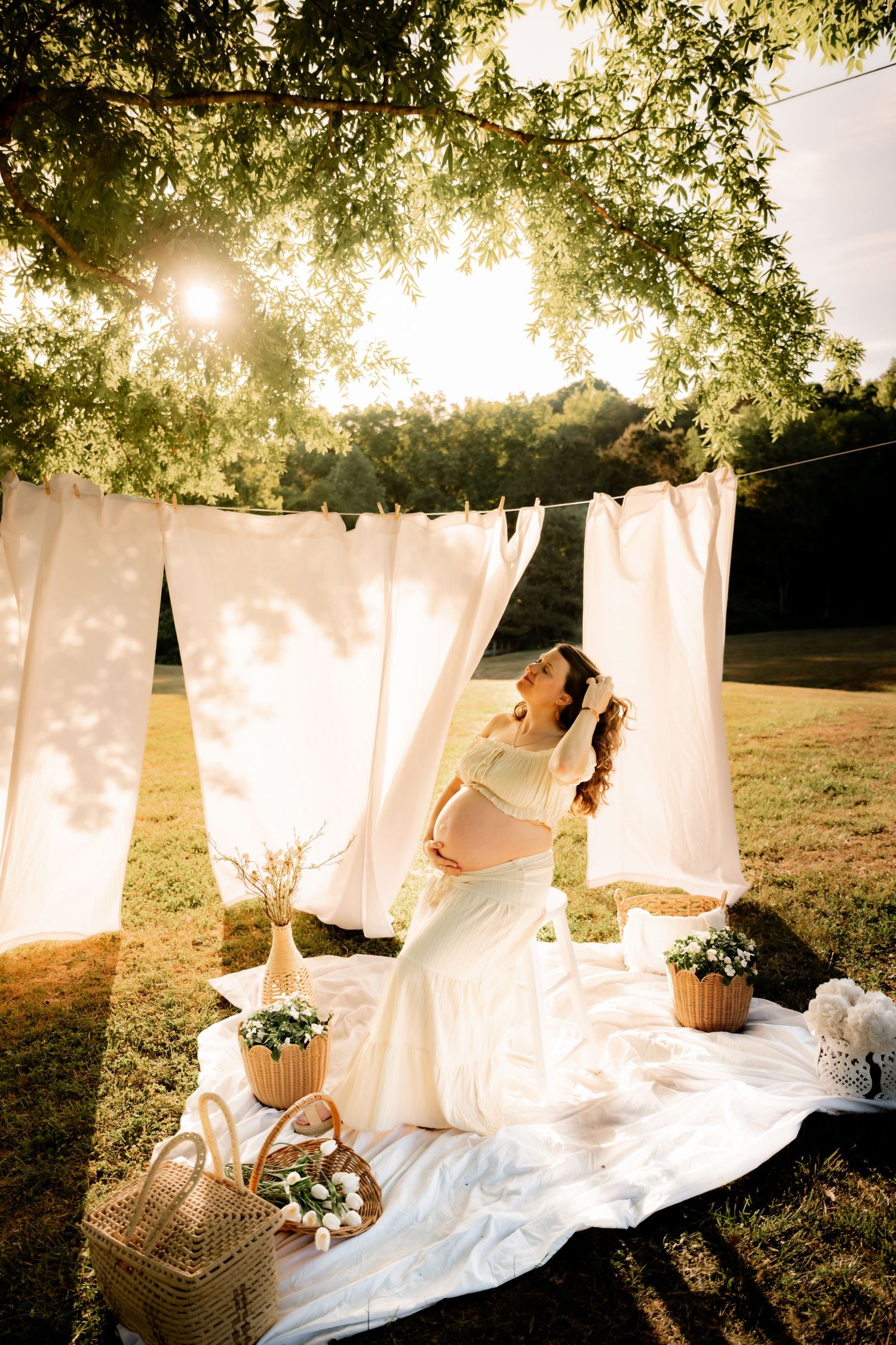 Pregnant woman in white dress poses outdoors at golden hour beneath white curtains and lush green trees.