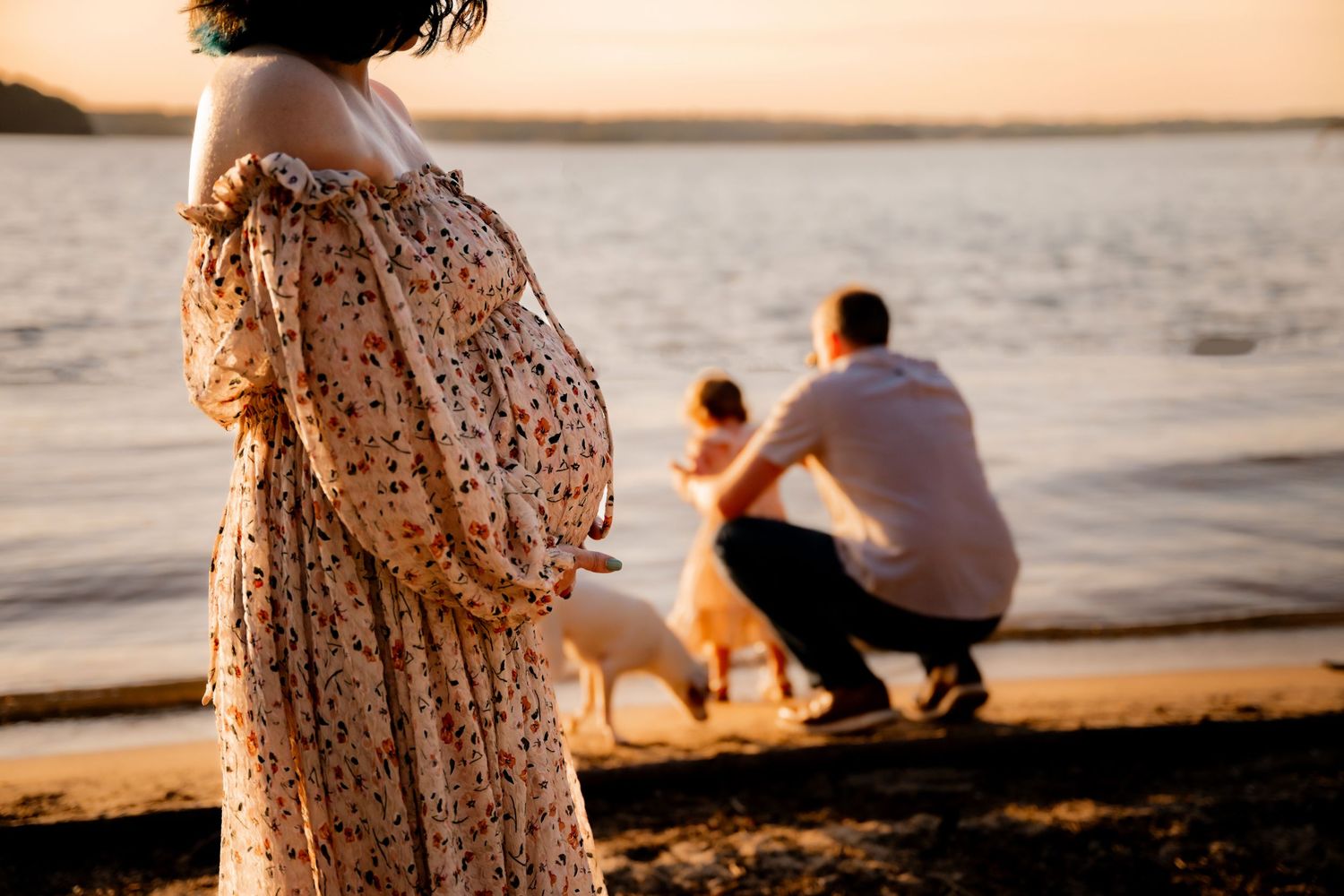 Pregnant woman in floral dress at sunset beach with partner and child petting a dog by the water.