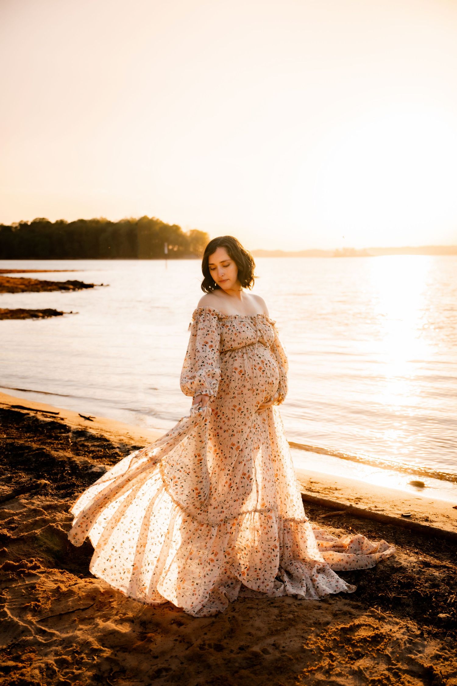 Pregnant woman in floral off-shoulder gown poses on sandy beach at golden sunset by calm water.